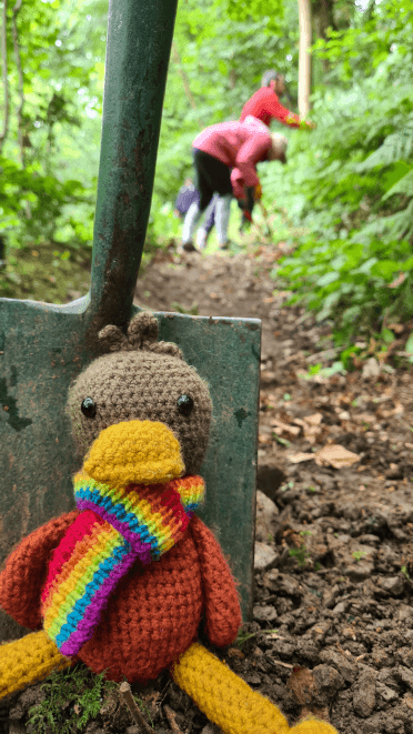 Croutons, the crocheted Contours Holidays mascot, sits at the base of a spade planted in the ground. In the background, Contours are hard at work volunteering to fix a damaged trail on conservation day.