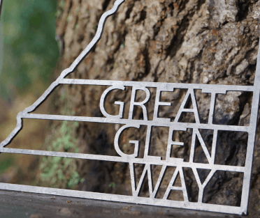 A winding line of metal copies the twists and turns of the Great Glen Way to create the main feature of this metal trail sign, complete with Great Glen Way in bold text beside. This one sits propped against an old tree.