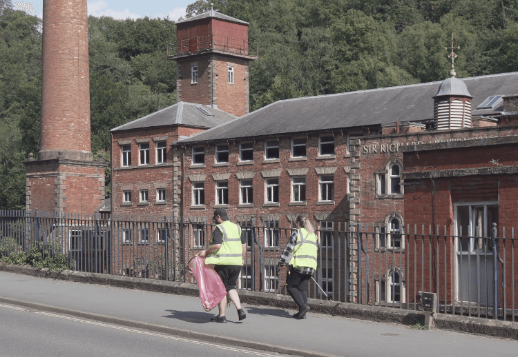Two members of the litter-picking team walk pass Masson Mill, a large, historic red-brick construction on the side of the A6.