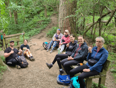 The Contours team take a snack break, sitting on the ground and a bench in the woods beside the Goose Nest Woods sign.